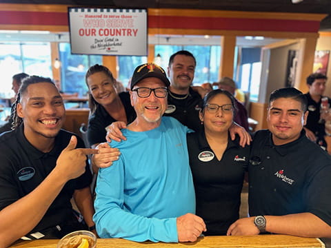 Applebee’s staff pose with a veteran in a blue shirt and cap seated at a table inside the restaurant